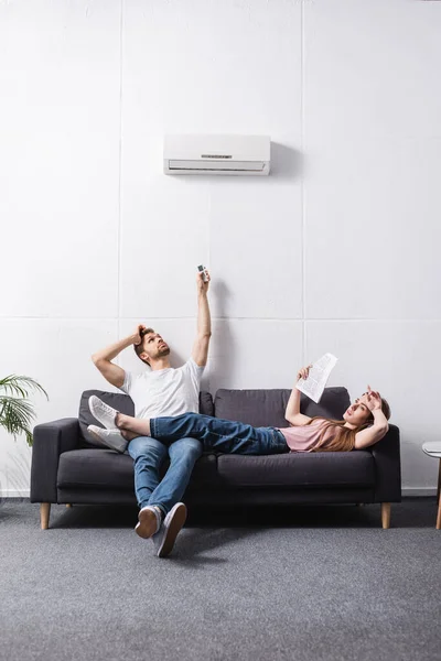 Jeune couple fatigué avec télécommande souffrant de chaleur à la maison avec climatiseur cassé — Photo de stock