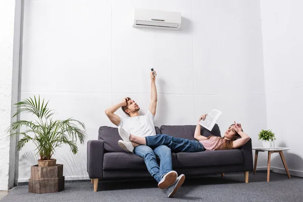 Frustrada pareja joven con mando a distancia que sufre de calor en casa con el aire acondicionado roto - foto de stock