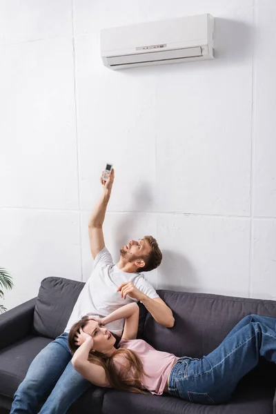 Frustrada pareja joven con mando a distancia que sufre de calor en casa con el aire acondicionado roto - foto de stock