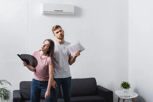 Casal exausto com ventilador de mão e jornal sofrendo de calor em casa com ar condicionado quebrado — Fotografia de Stock