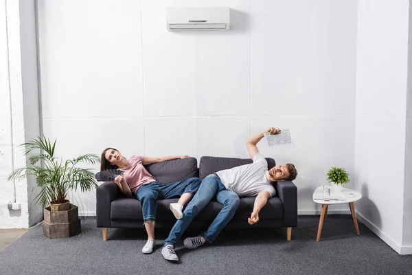 Couple triste avec ventilateur à main et journal souffrant de chaleur à la maison avec climatiseur cassé — Photo de stock