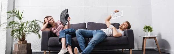 Jeune couple avec ventilateur à main et journal souffrant de chaleur à la maison, orientation panoramique — Photo de stock