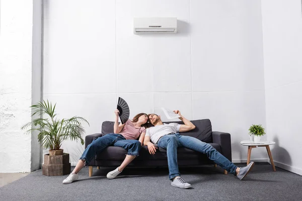 Young couple with hand fan and newspaper suffering from heat at home with broken air conditioner — Stock Photo