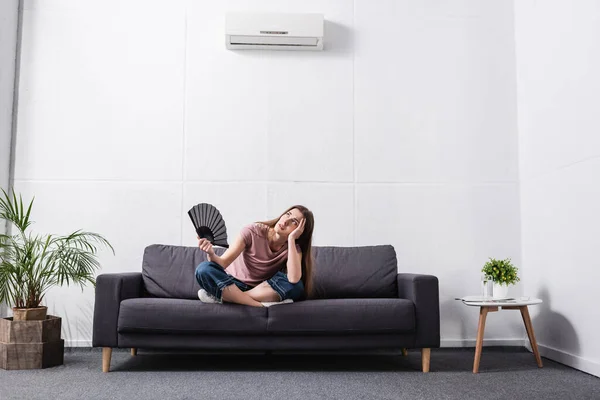 Mujer joven confundida que sufre de calor y el uso de ventilador de mano en casa con acondicionador roto - foto de stock