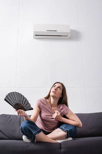 Jovem que sofre de calor e usando ventilador de mão em casa com condicionador quebrado — Fotografia de Stock
