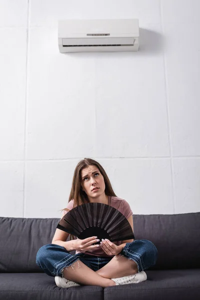 Upset girl suffering from heat and using hand fan at home with broken conditioner — Stock Photo