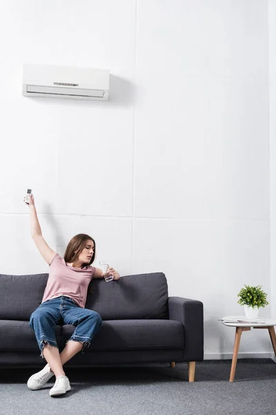 Aufgebrachte junge Frau hält Glas Wasser in der Hand, während sie unter Hitze leidet und versucht, Klimaanlage einzuschalten — Stockfoto