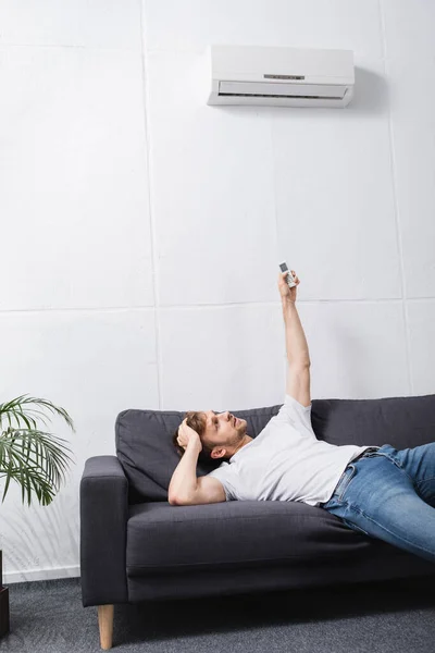 Homem confuso segurando controlador remoto e sofrendo de calor com ar condicionado quebrado em casa — Fotografia de Stock