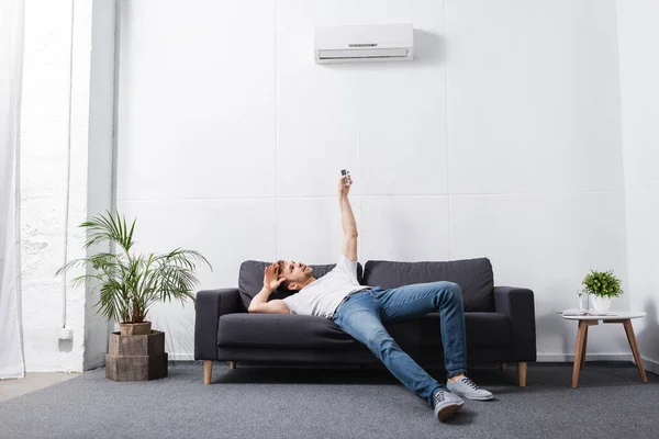Exhausted man holding remote controller and suffering from heat with broken air conditioner at home — Stock Photo