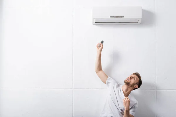 Смущенный человек держит пульт дистанционного управления и страдает от жары со сломанным кондиционером воздуха дома — стоковое фото