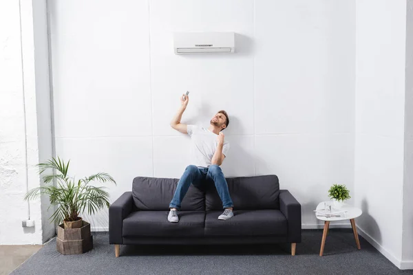 Homem cansado segurando controlador remoto e sofrendo de calor com ar condicionado quebrado em casa — Fotografia de Stock