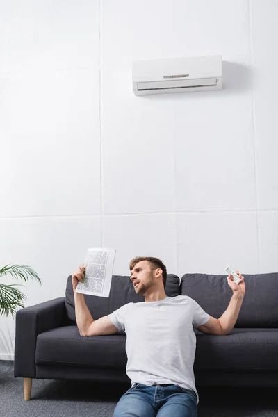 Jovem triste que sofre de calor ao usar o jornal como ventilador de mão em casa com ar condicionado quebrado — Fotografia de Stock