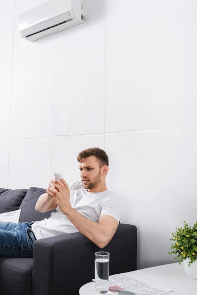 Uomo scettico cercando di accendere condizionatore con telecomando a casa — Foto stock