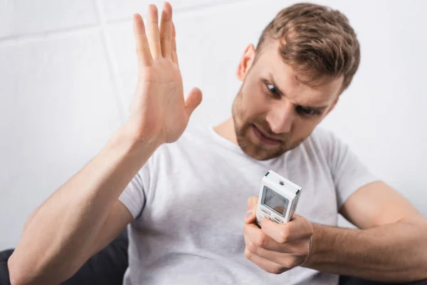 Irritated man trying to switch on air conditioner with broken remote controller at home — Stock Photo