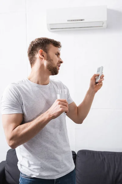 Sad man holding glass of water while trying to switch on air conditioner with broken remote controller at home — Stock Photo