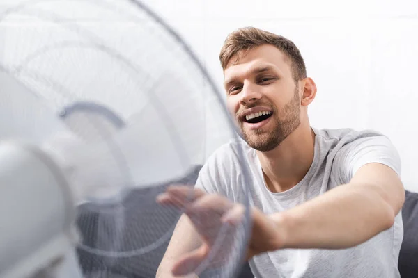 Красивый позитивный мужчина чувствует себя комфортно с электрическим вентилятором дома в летнюю жару — стоковое фото