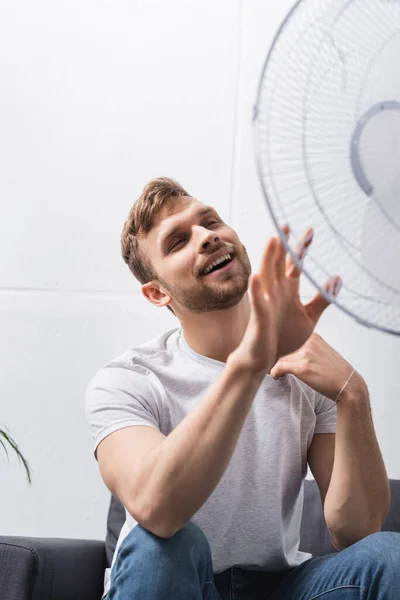 Hombre positivo sentirse cómodo con ventilador eléctrico en casa durante el calor del verano - foto de stock