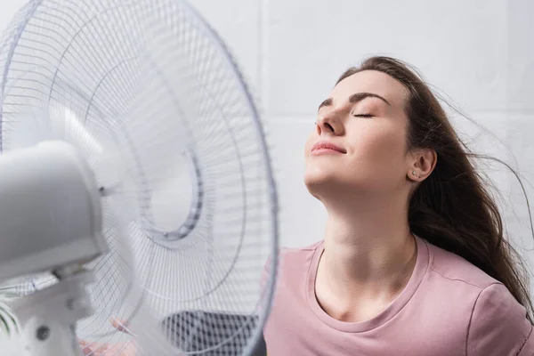 Atractiva chica positiva sentirse cómodo con ventilador eléctrico en casa durante el calor del verano - foto de stock