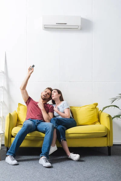 Casal confuso com controlador remoto sofrendo de calor em casa com ar condicionado quebrado — Fotografia de Stock