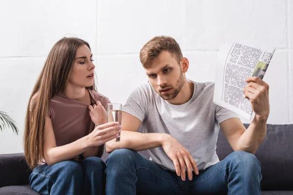 Young couple suffering from heat at home with newspaper and glass of water — Stock Photo