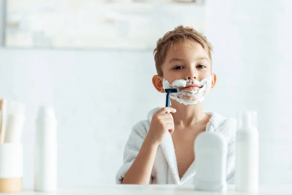 Enfoque selectivo de lindo chico afeitándose en el baño cerca de artículos de tocador - foto de stock