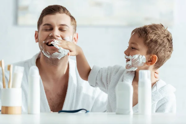 Enfoque selectivo de niño sonriente aplicando espuma de afeitar en la cara de los padres - foto de stock