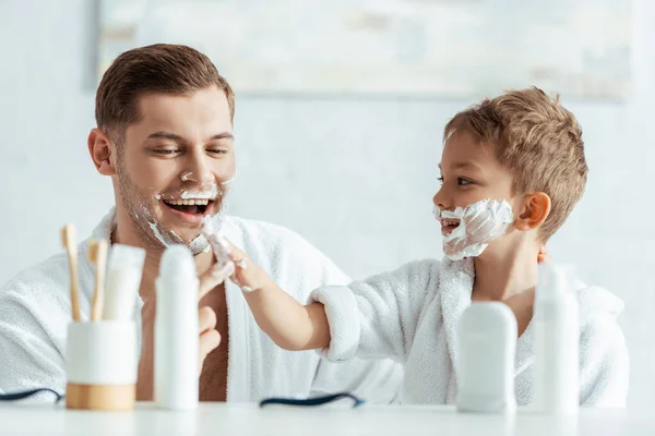 Selective focus of smiling boy applying shaving foam on face of cheerful father — Stock Photo