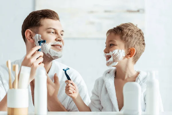 Selektiver Fokus eines lächelnden Jungen mit Schaum im Gesicht, der seinen rasierenden Vater betrachtet — Stockfoto