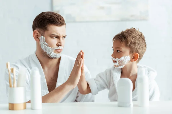 Enfoque selectivo de padre e hijo serios con espuma de afeitar en las caras dando alta cinco - foto de stock