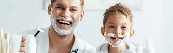 Horizontal image of happy father and son with shaving foam on faces holding shaving razors — Stock Photo