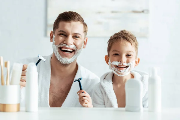 Selektiver Fokus von fröhlichen Vater und Sohn mit Rasierschaum auf Gesichtern, die Rasiermesser halten — Stockfoto