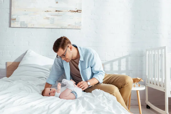 Glücklicher junger Vater berührt niedlichen Jungen, der im Strampler auf dem Bett liegt — Stockfoto