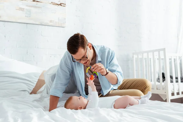 Happy young man playing with baby rattle over cute infant lying in bed — Stock Photo