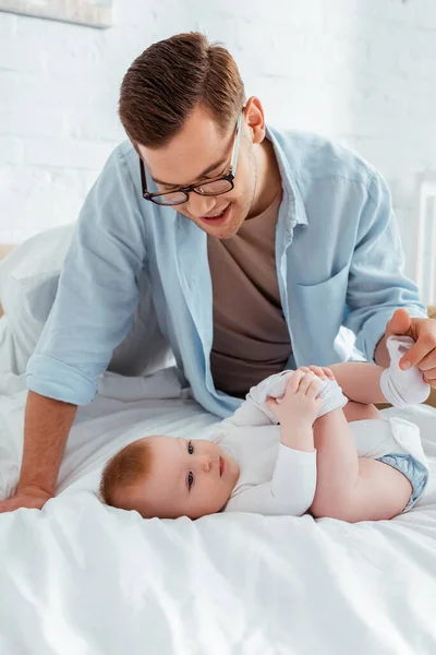 Glücklicher junger Vater berührt Bein seines entzückenden kleinen Sohnes, der im Bett liegt — Stockfoto