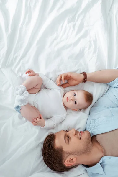 Top view of happy young man touching adorable baby boy while lying near him in bed — Stock Photo