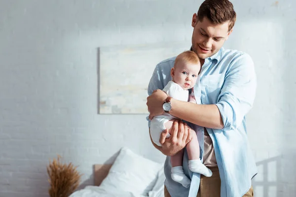 Feliz joven padre sosteniendo y abrazando adorable pequeño hijo en el dormitorio - foto de stock