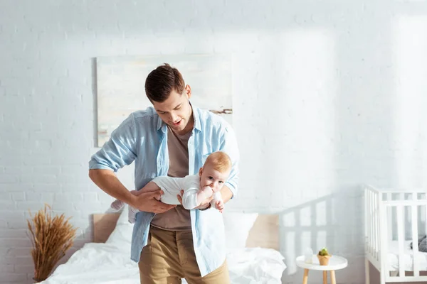 Joven padre sosteniendo lindo bebé niño en mameluco mientras de pie en el dormitorio - foto de stock