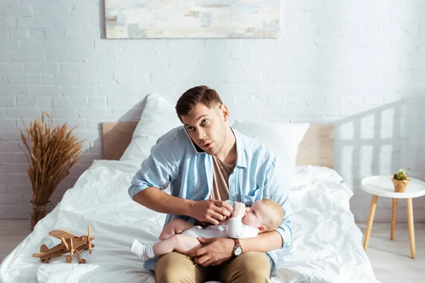 Attentive man talking on smartphone while feeding cute little son from baby bottle — Stock Photo