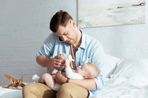 Young father feeding baby boy from baby bottle while talking on smartphone — Stock Photo