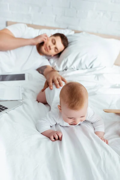 Selective focus of man talking on smartphone with closed eyes while touching baby boy crawling on bed — Stock Photo