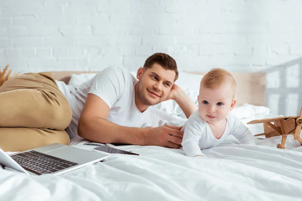 Счастливый мужчина, лежащий на кровати рядом с обожаемым сыном, ноутбуком и игрушечным самолетом — стоковое фото