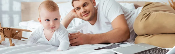 Панорамная ориентация счастливого отца и милого младенца на кровати рядом со смартфоном на игрушечном самолете — стоковое фото