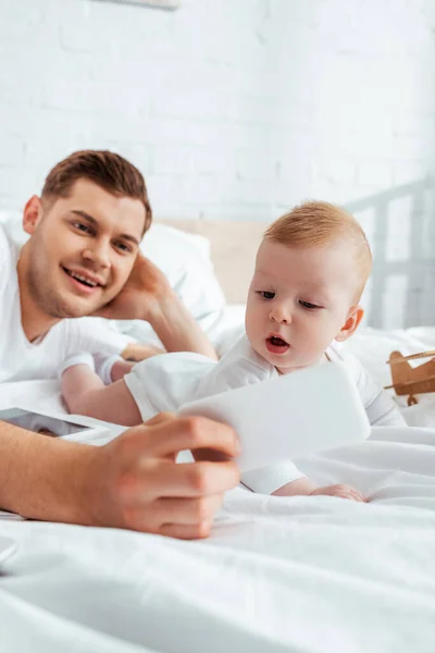 Selective focus of smiling father taking selfie on smartphone with adorable little son — Stock Photo