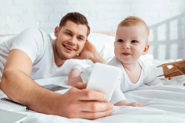 Selective focus of happy father taking selfie on smartphone with smiling baby boy — Stock Photo