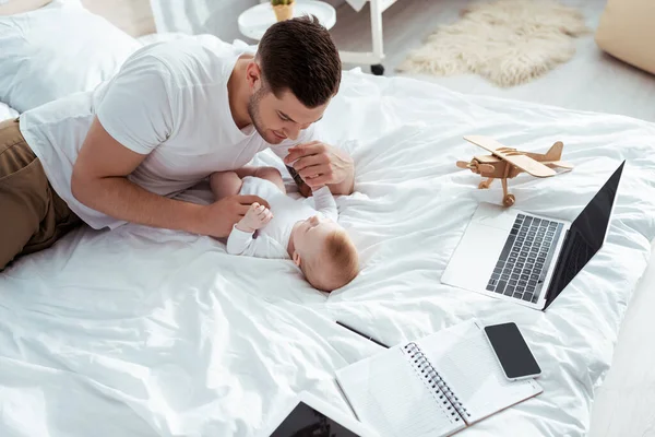 Young father playing with baby boy near gadgets, notebook and toy plane on bed — Stock Photo