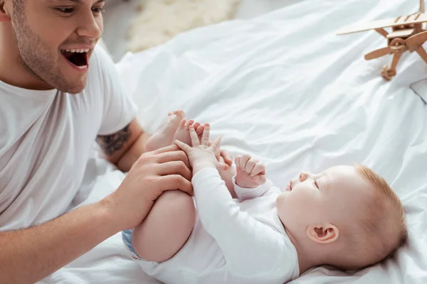 Glücklicher, aufgeregter Vater berührt Beine seines entzückenden Sohnes, der im Bett liegt — Stockfoto