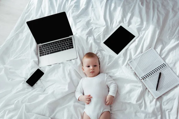Draufsicht des niedlichen Säuglings auf dem Bett liegend in der Nähe von Gadgets und Notizbuch mit Stift — Stockfoto
