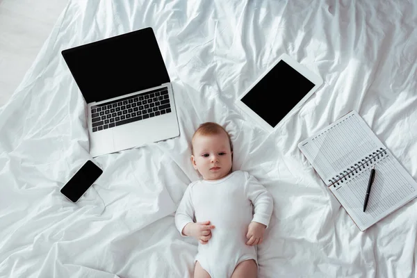 Vista dall'alto del bambino carino sdraiato sul letto vicino a gadget e notebook con penna — Foto stock