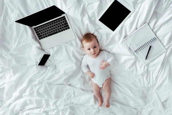 Vue de dessus de l'adorable bébé garçon couché sur le lit près de gadgets et ordinateur portable avec stylo — Photo de stock