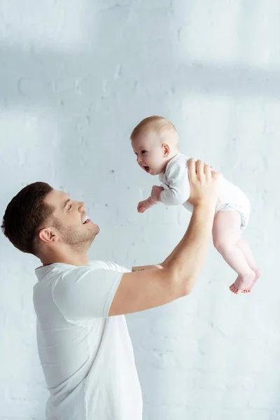 Alegre padre joven sosteniendo lindo bebé en manos extendidas en el dormitorio - foto de stock